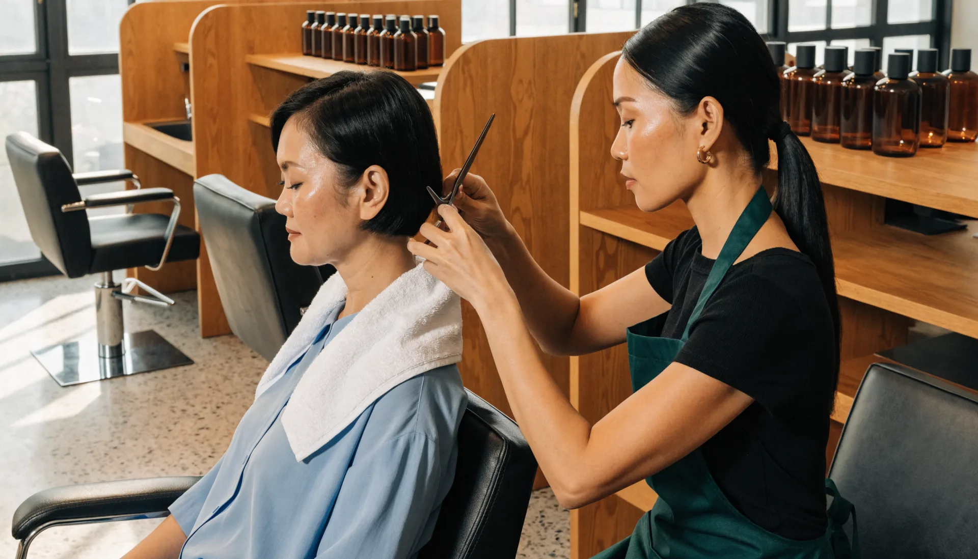 The stylist trims a client’s bob at her station, a shelf of hair products behind them.
