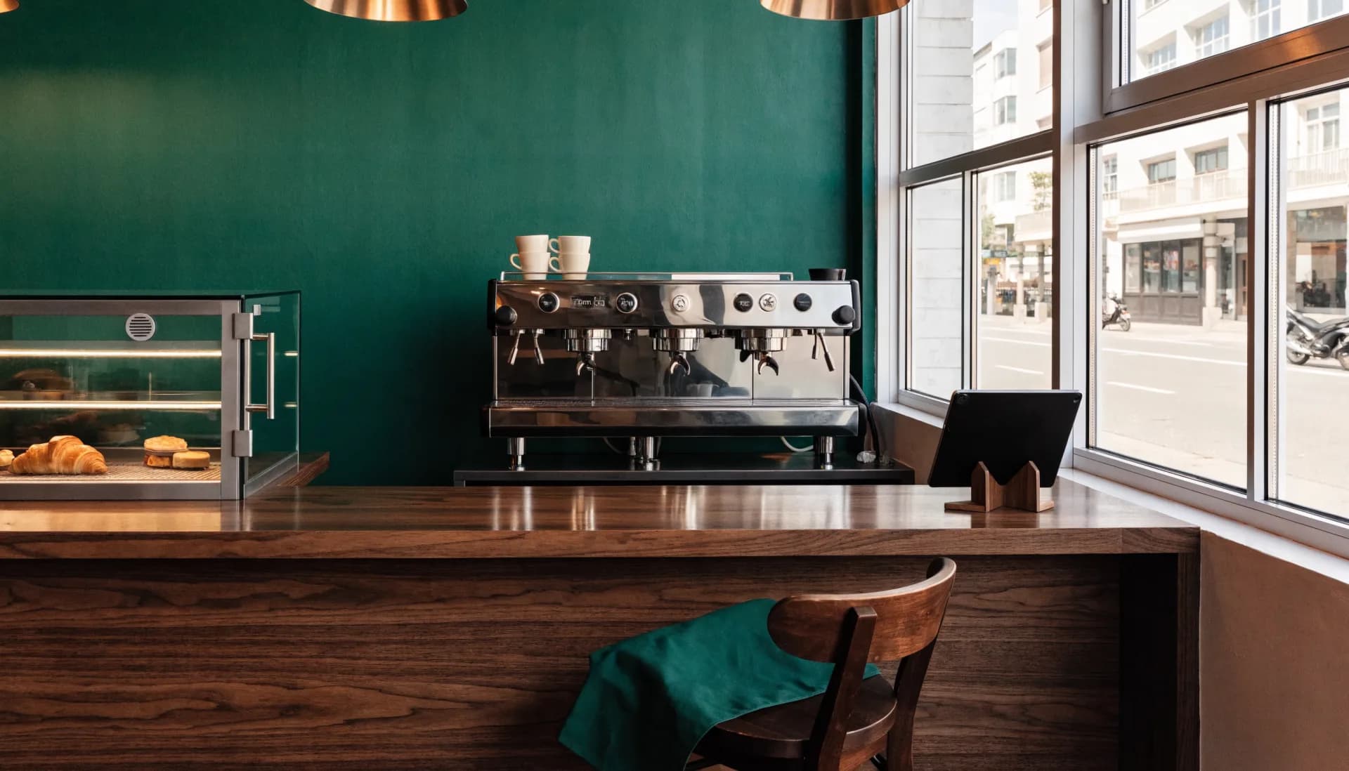 The bar at rest — walnut counter wiped clean, cups stacked on the chrome machine, the tablet on its stand by the window, a green apron folded over the chair.