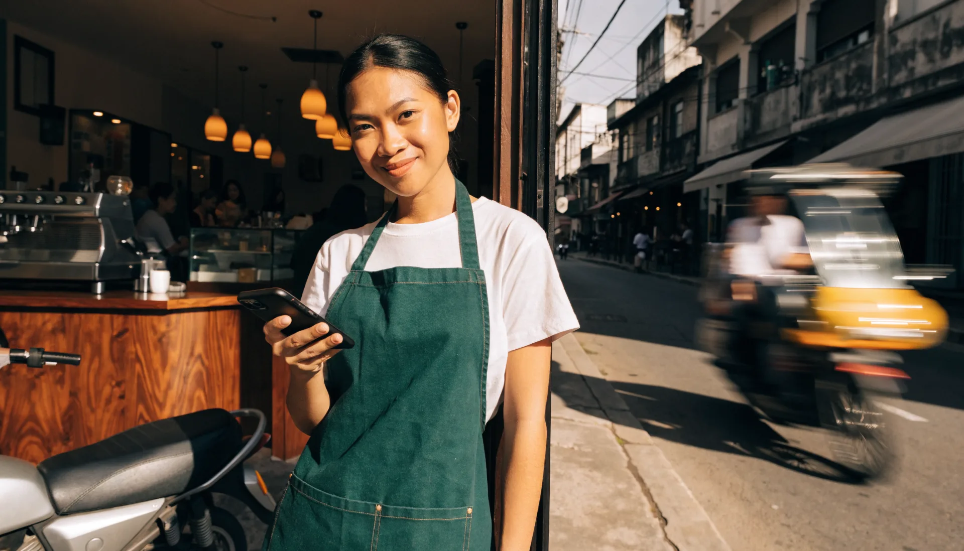 The owner in the doorway of her shop with her phone, the coffeehouse busy behind her and a tricycle passing on the street.
