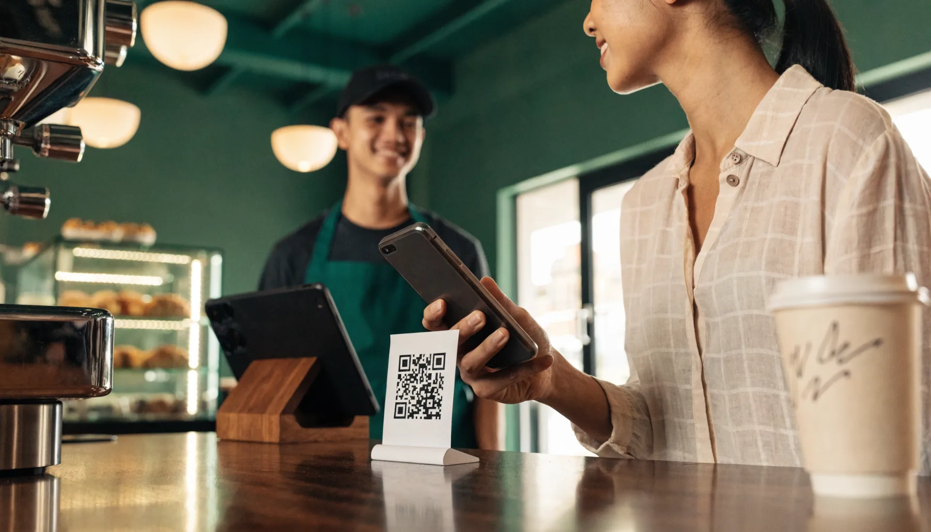 A customer holds her phone over the QR standee on the counter while the cashier smiles behind the register tablet.