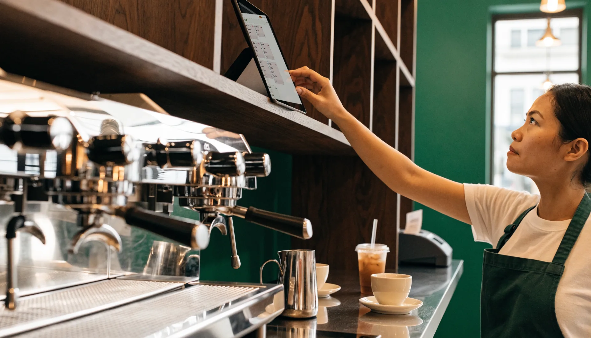 A barista reaches up to the kitchen display tablet on the shelf above the espresso machine, where the open orders are listed as tickets.