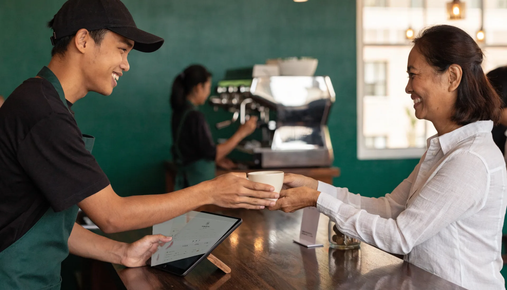 A barista hands a cup across the counter to a smiling regular while the register tablet beside him shows her customer profile.