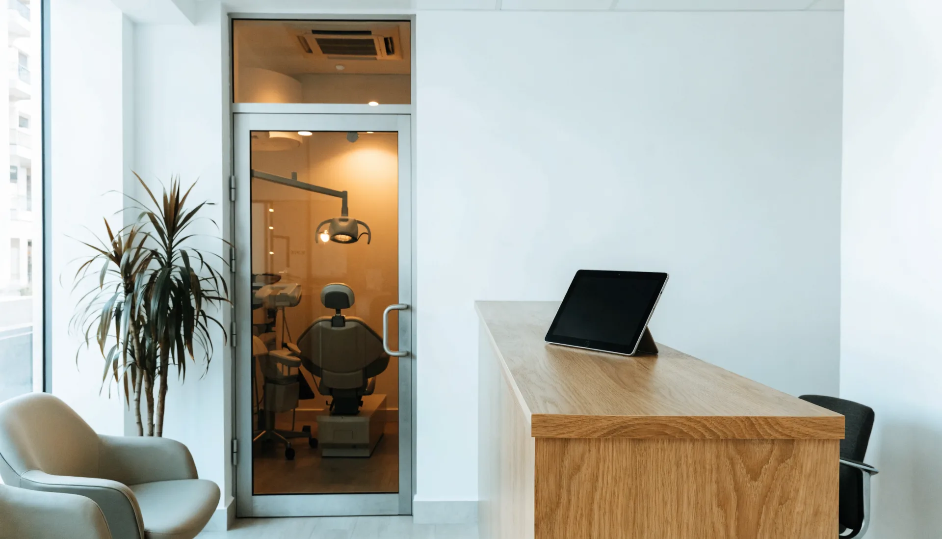 The clinic at rest before opening — a quiet waiting area, the treatment room through the glass door, the tablet on the front desk.
