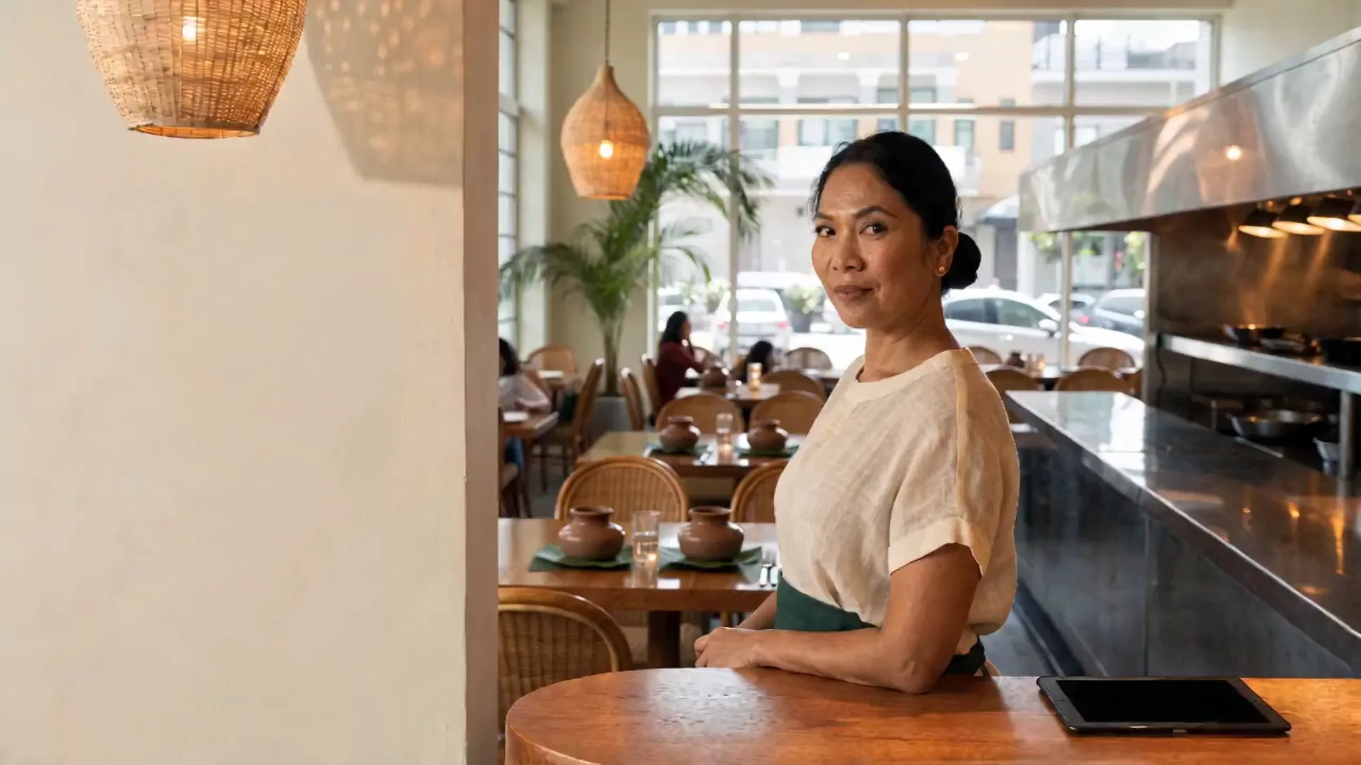 The owner at the host counter of her Filipino restaurant, calm and smiling, with diners at lunch and the open kitchen behind her.