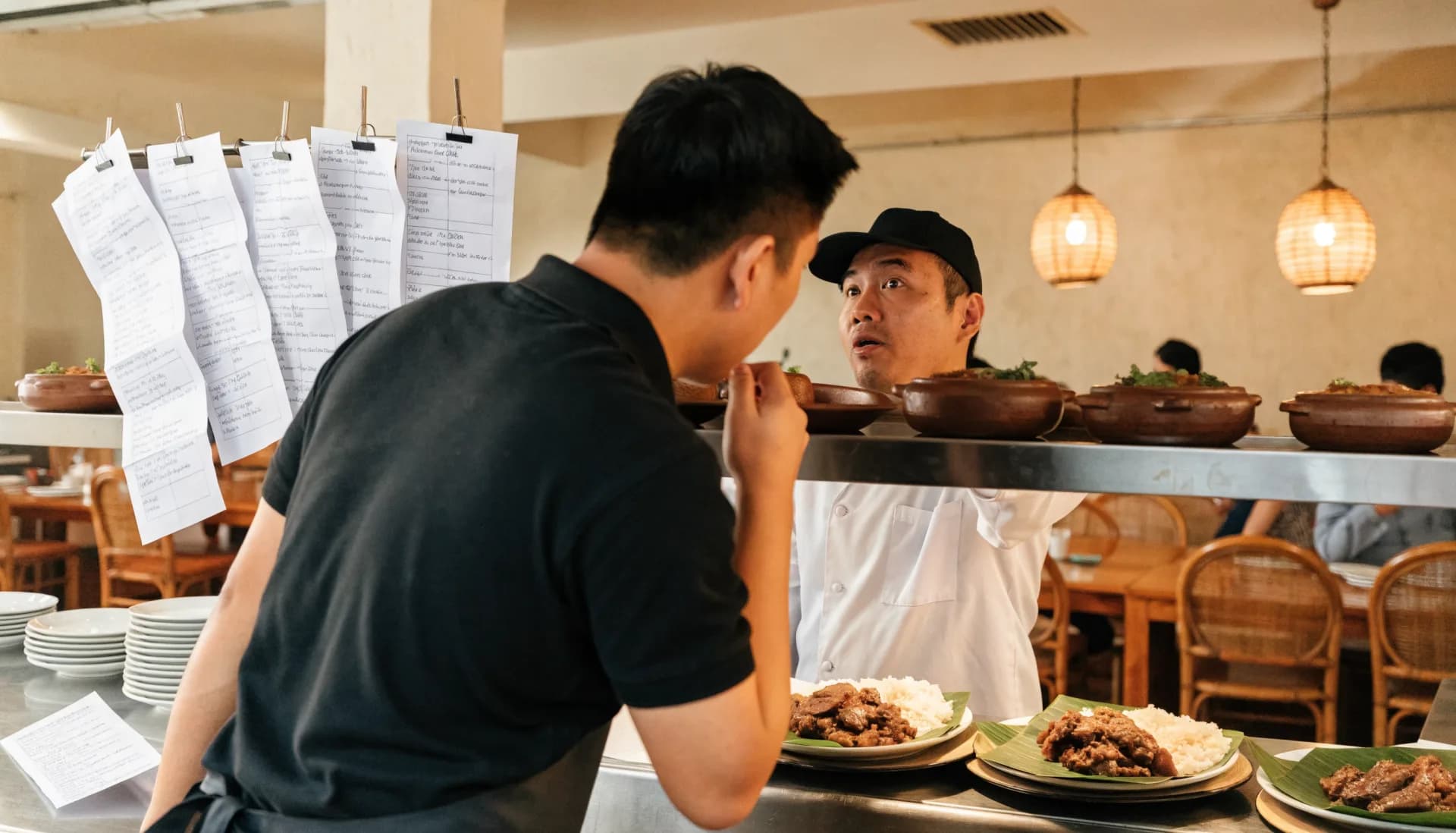 The lunch rush without a system — handwritten order slips pile up on the kitchen rail while a server leans in to repeat an order and the cook looks up, unsure.